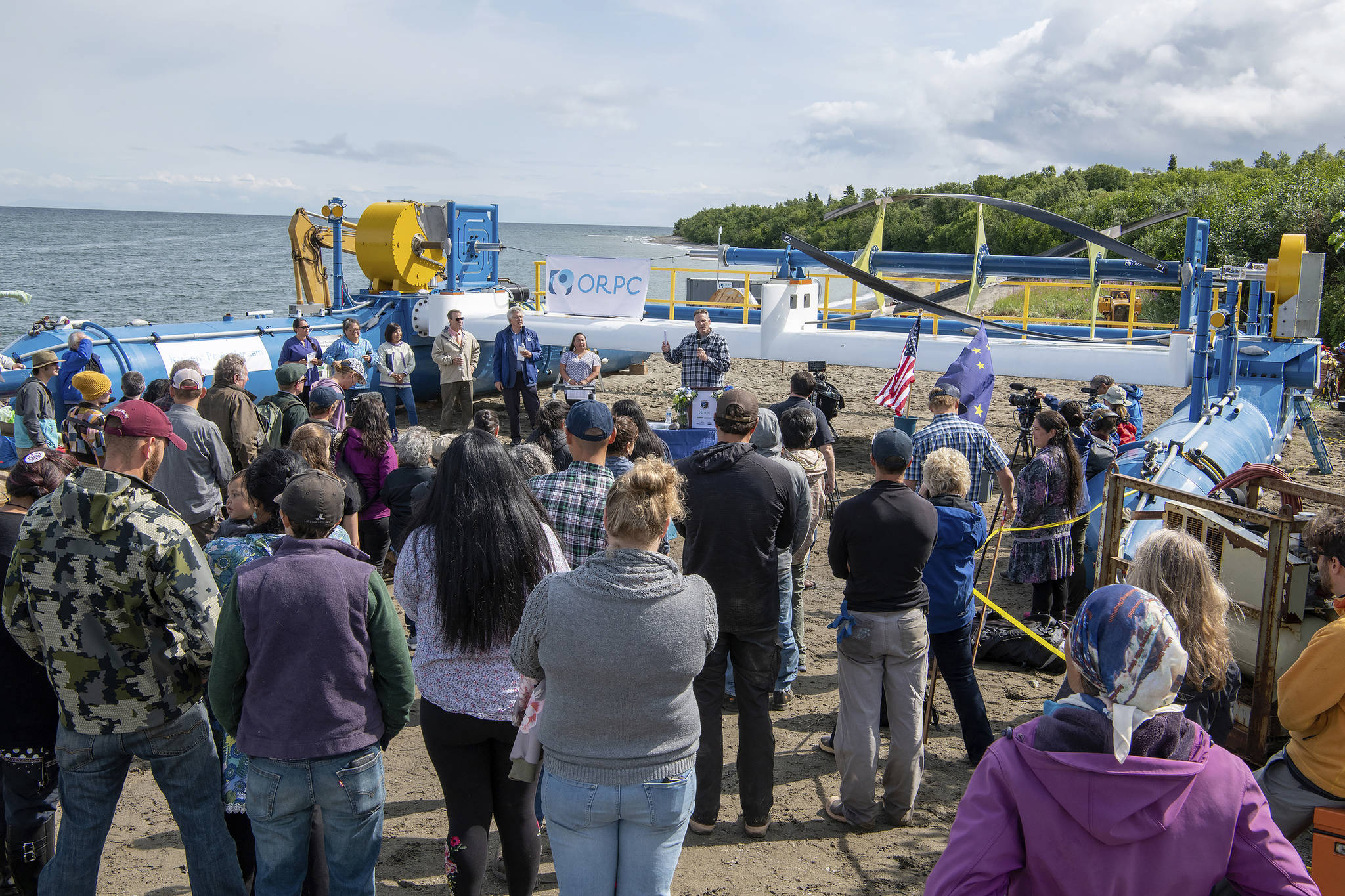 In this Tuesday, July 16, 2019, photo released by the Alaska Governor’s office, Gov. Mike Dunleavy, center rear, speaks in front of a Riv-Gen Power System turbine on the bank of the Kvichak River in Igiugig, Alaska. A tiny Alaska Native village is adopting an emerging technology to transform the power of a local river into a renewable energy source. (Austin McDaniel/Alaska Governor’s Office via AP)