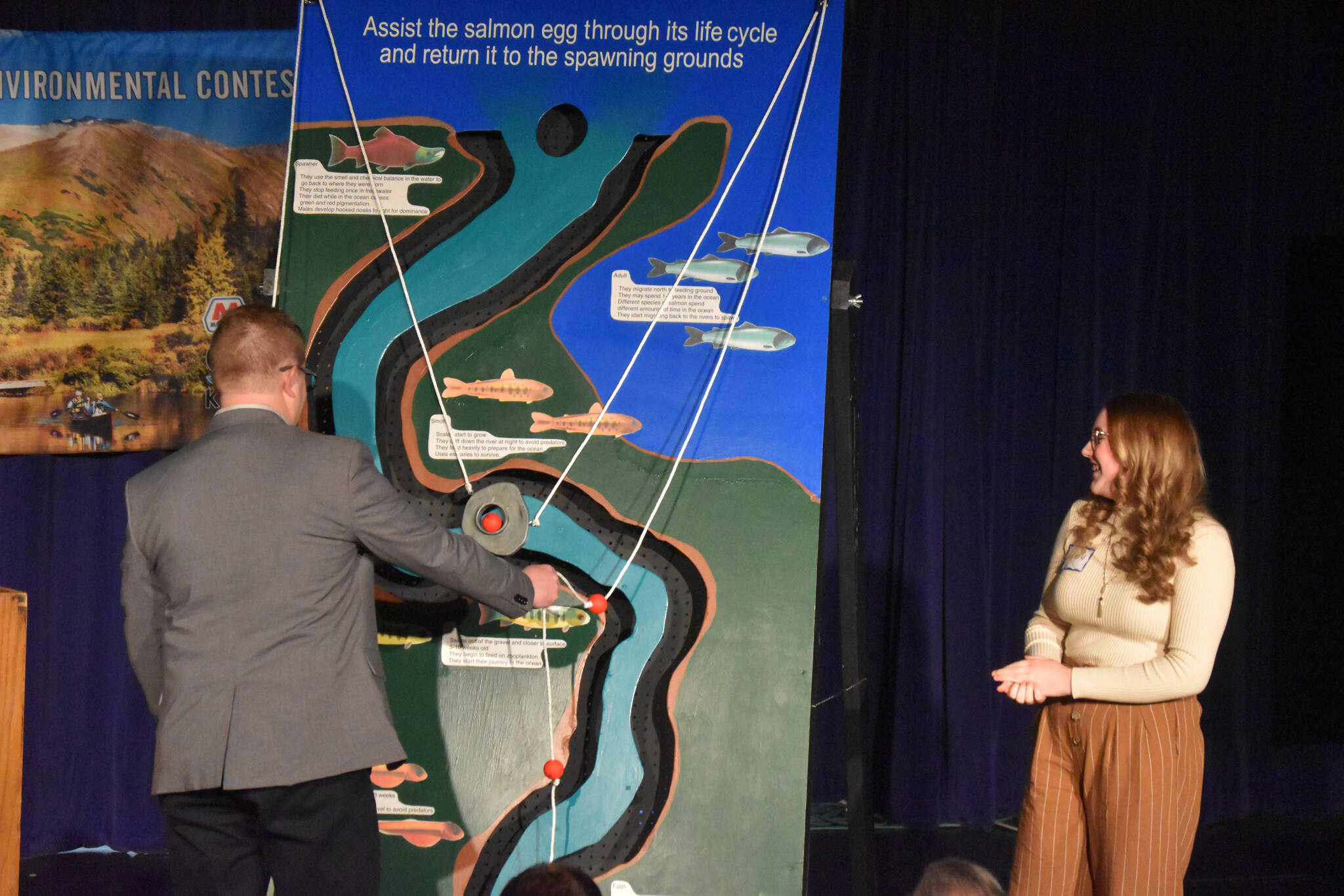 Soldotna High School student Emerson Kapp, second-place winner, is joined on stage by judge Bruce Jackman as she presents the Kenai Peninsula Maze Board during the 33rd Caring for the Kenai competition at Kenai Central High School in Kenai, Alaska on Thursday, April 20, 2023.