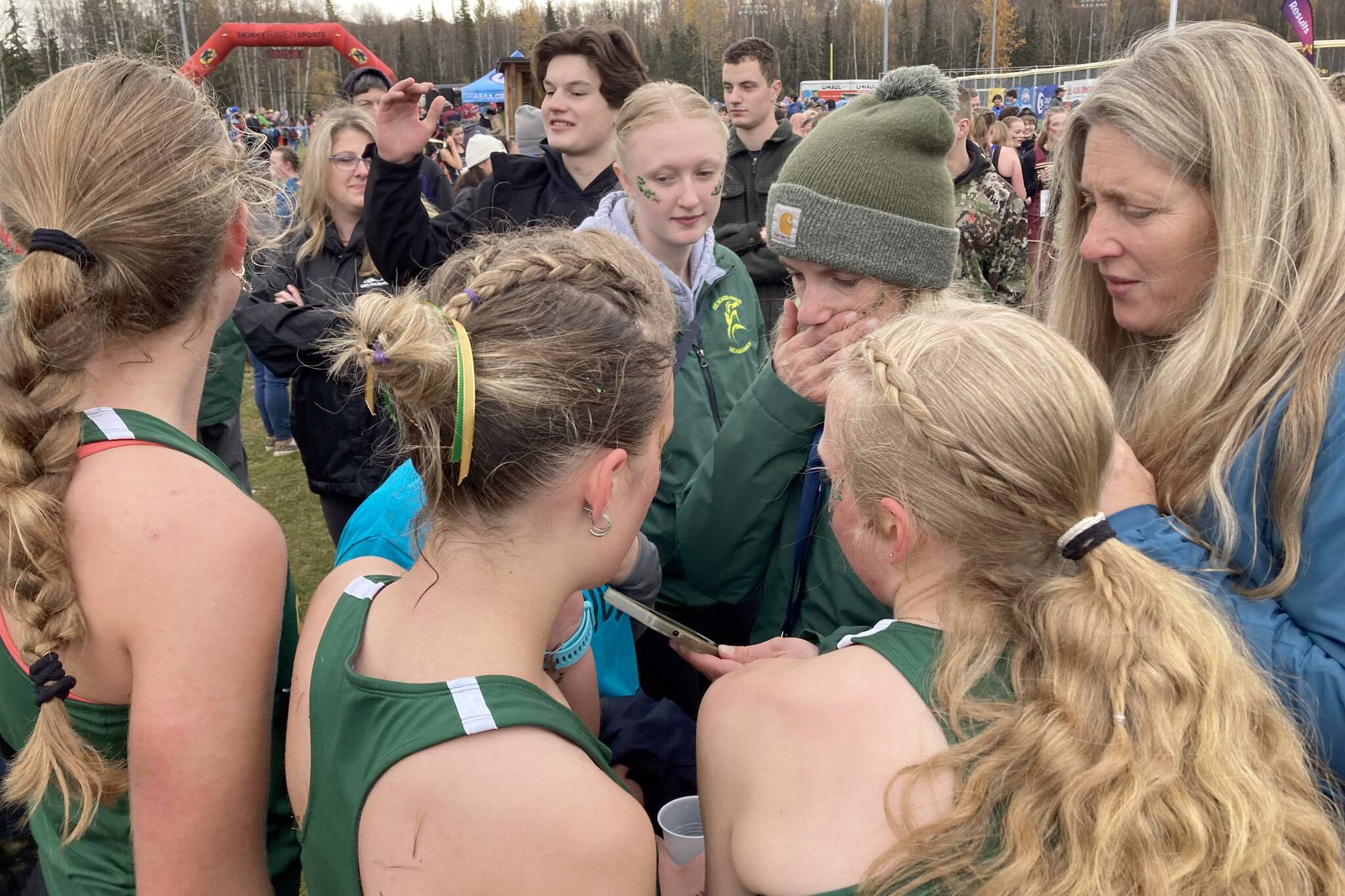 Seward cross-country head coach Rachel Dow looks at results on her phone while her team huddles around her at the state cross-country running meet on Saturday, Oct. 5, 2024, at Bartlett High School in Anchorage, Alaska. The Seahawks won a third straight Division II girls title. (Photo by Jeff Helminiak/Peninsula Clarion)