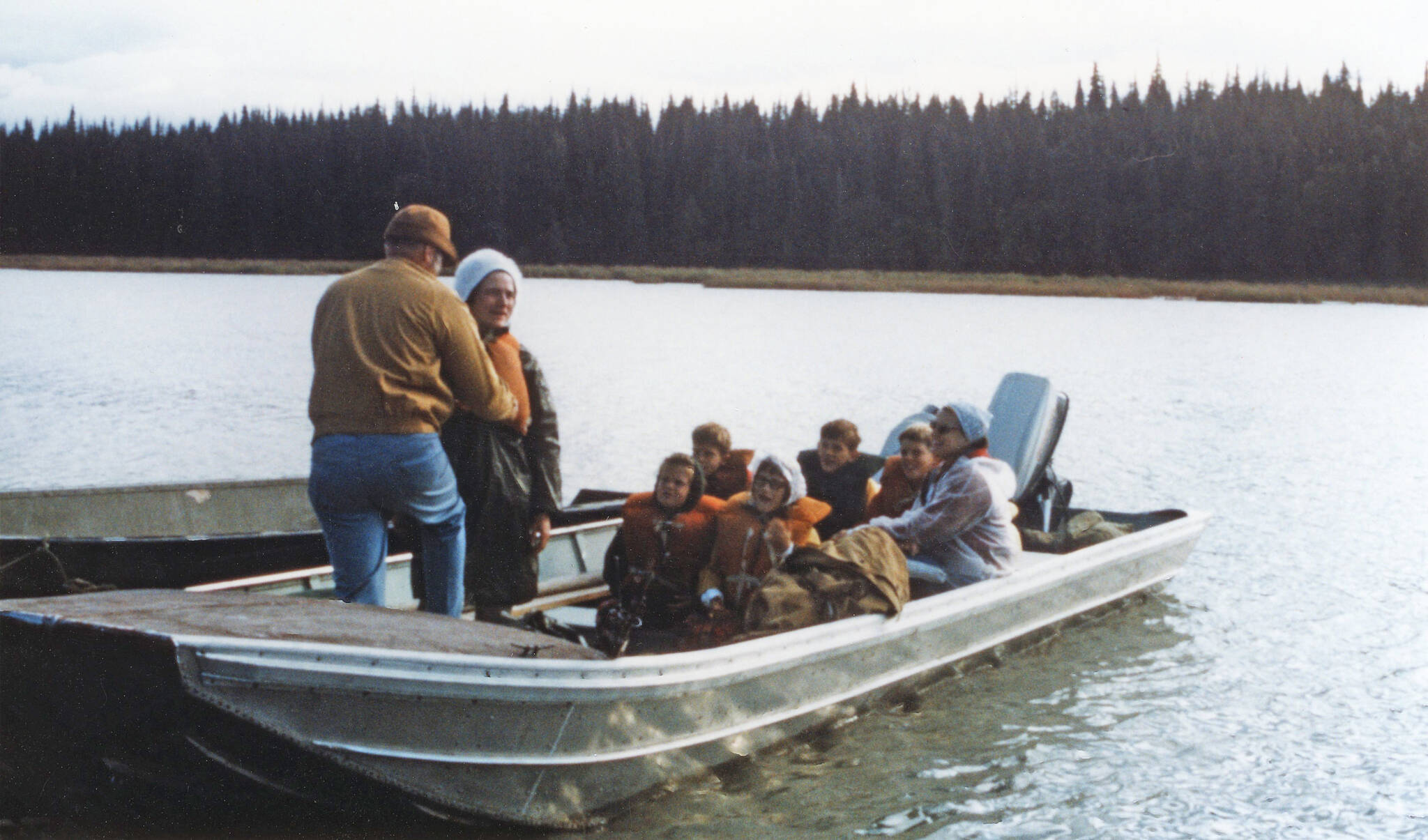 Jane Fair (standing, wearing white hat) receives help with her life jacket from Ron Hauswald prior to the Fair and Hauswald families embarking on an August 1970 cruise with Phil Ames on Tustumena Lake. Although conditions were favorable at first, the group soon encountered a storm that forced them ashore. (Photo courtesy of the Fair Family Collection)