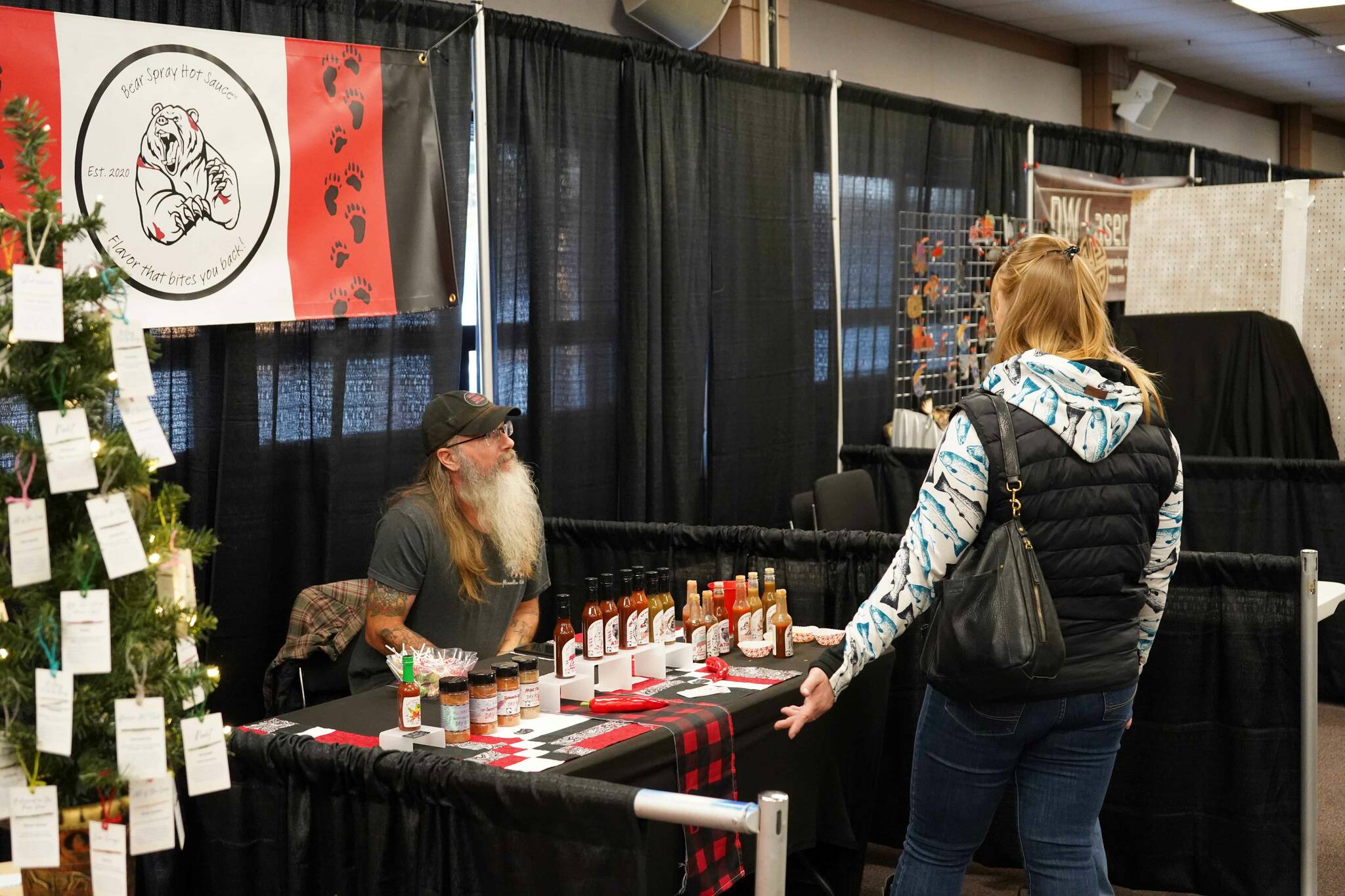 Terry Holmes, of Bear Spray Hot Sauce, chats with shoppers at the 33rd Annual Holiday Bazaar at the Soldotna Regional Sports Complex in Soldotna, Alaska, on Friday, Nov. 22, 2024. (Jonas Oyoumick/Peninsula Clarion)